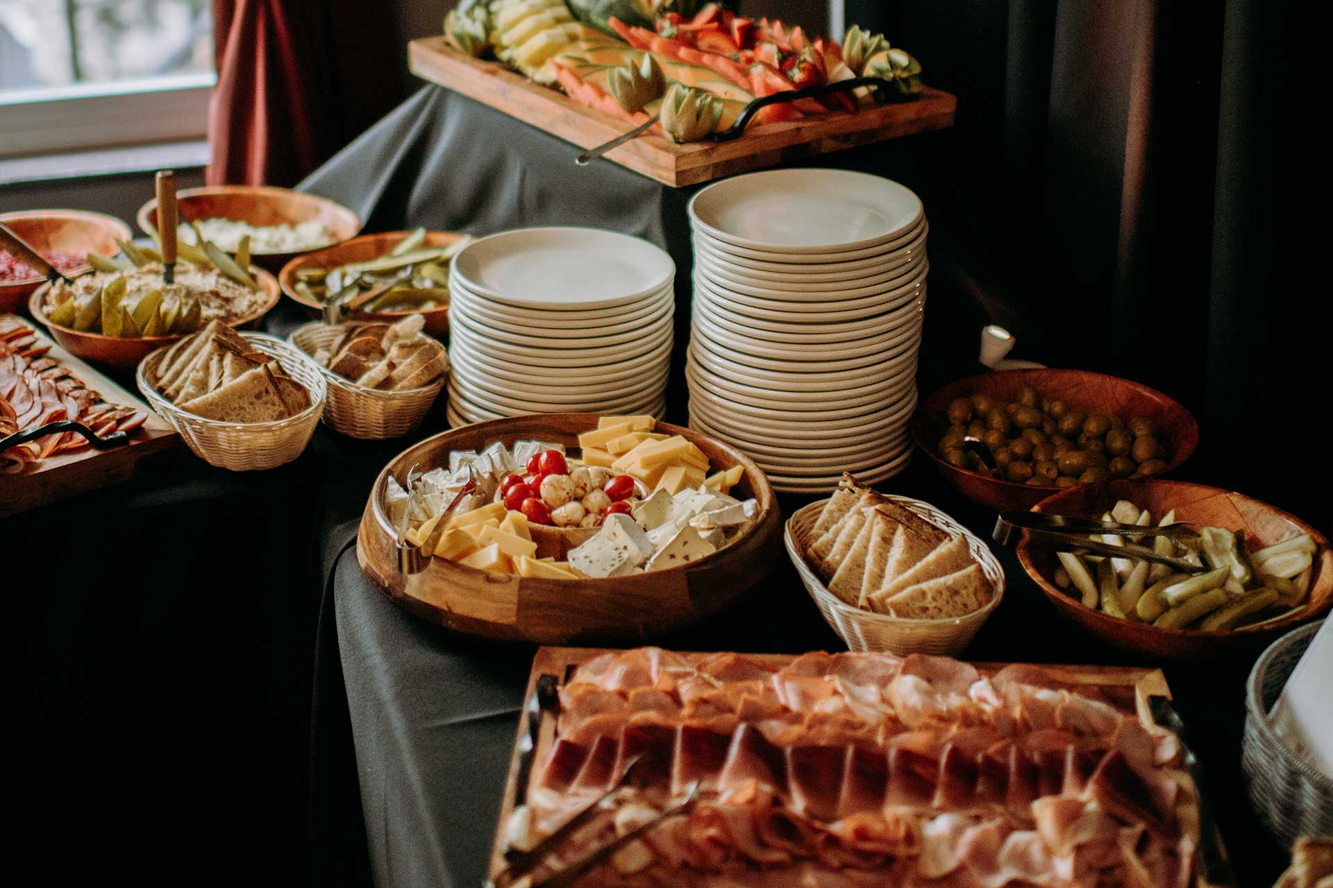 sebis-kitchen-holiday-menu-intro2 Buffet table with assorted cheeses, olives, sliced meats, and bread. Plates are stacked for serving at an event or wedding catering.