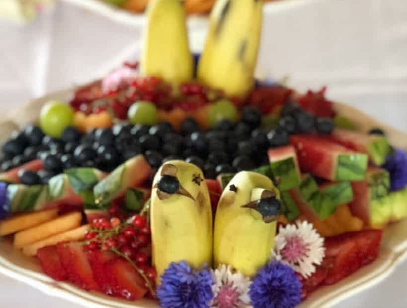 Decorative fruit platter with bananas shaped like birds, surrounded by berries, grapes, and colorful flowers, ideal for events.