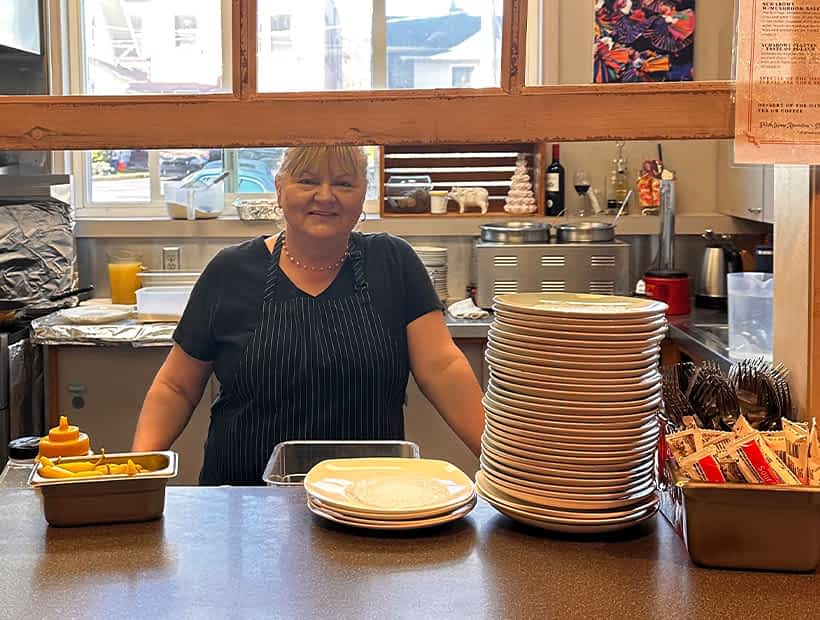 Chef Kamila Kanczugowski in a kitchen with a sign reading This kitchen is seasoned with love, standing behind stacked plates and cutlery at a counter.